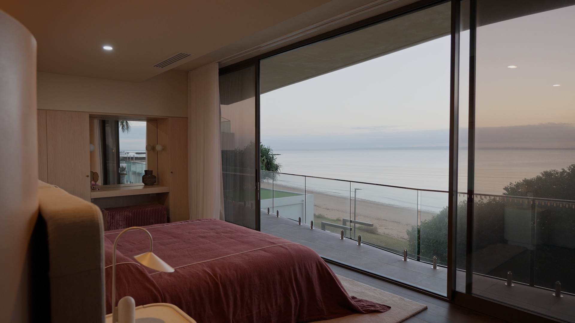 Bedroom opening onto a glass balcony above the beach, bay and horizon at first light