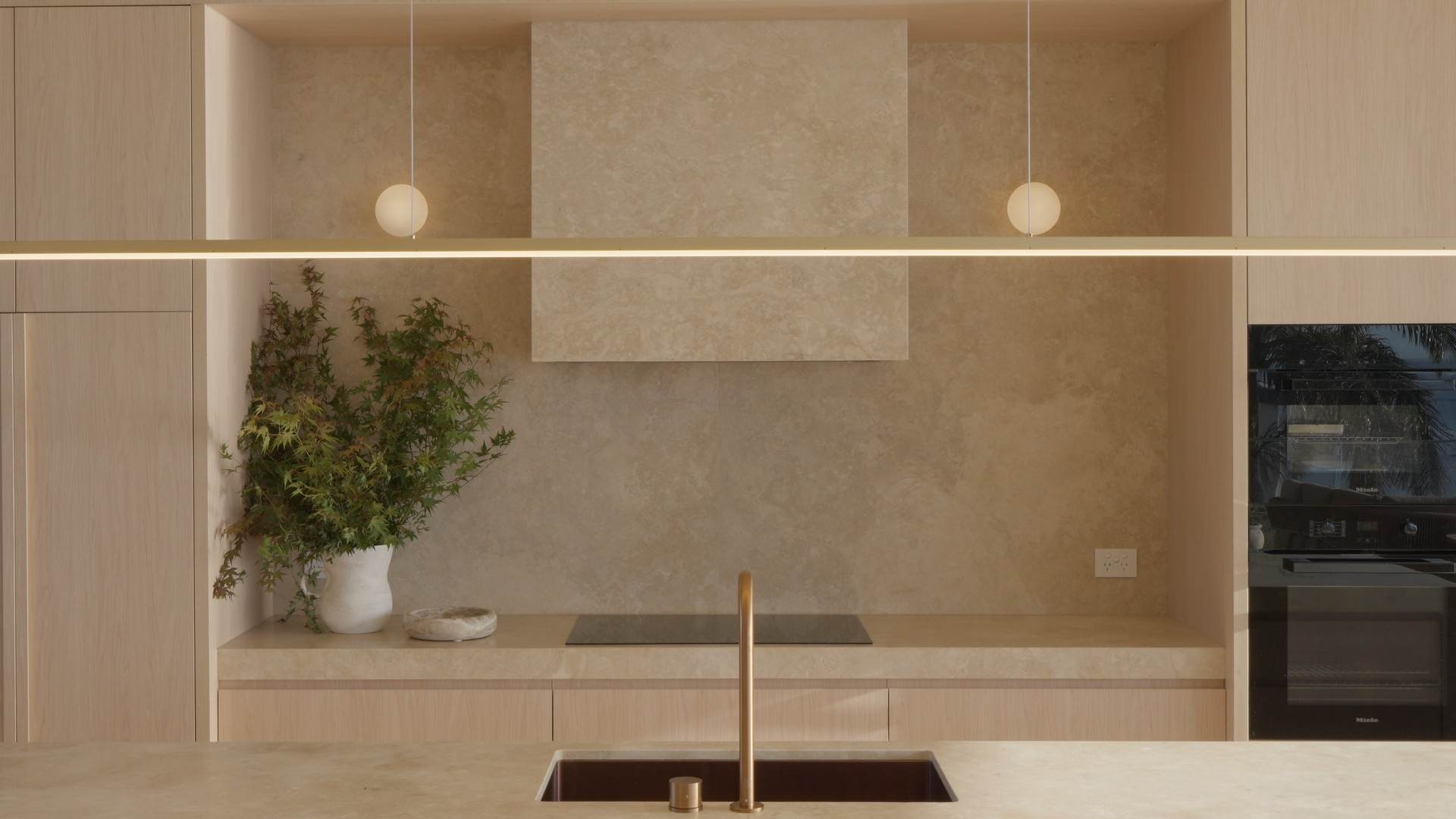 Travertine kitchen with oak joinery, brass tap and a linear pendant glowing across the frame